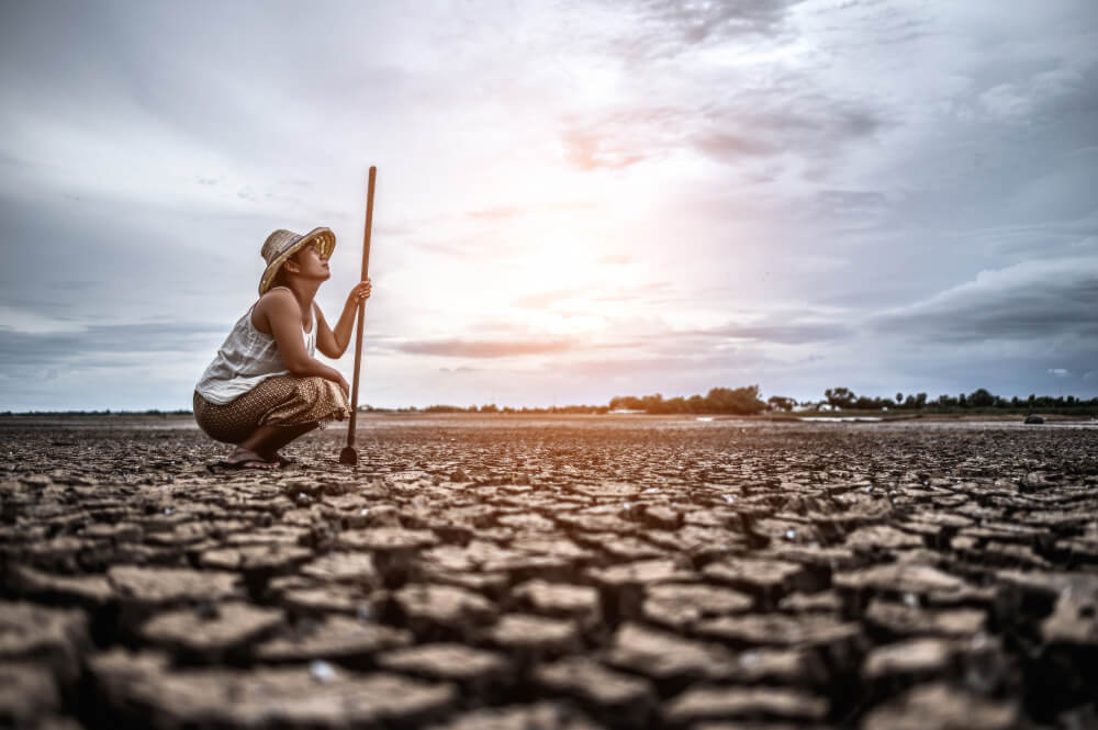 Imagem mostra mulher agricultora, agachada em solo seco, olhando para o céu.